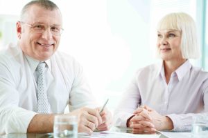 Portrait of smiling boss looking at camera at workplace with mature woman looking at him near by